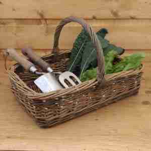 Golden brown rectangular willow garden trug with two green bands and a wrapped bow handle, with garden tools and fresh vegetables